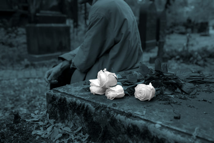 Eine Person sitzt vor einem Grabstein, auf dem weiße Rosen liegen – Symbol für die Bestattungsvorsorge von RIANEVA, die finanzielle Sicherheit und Entlastung im Trauerfall bietet.