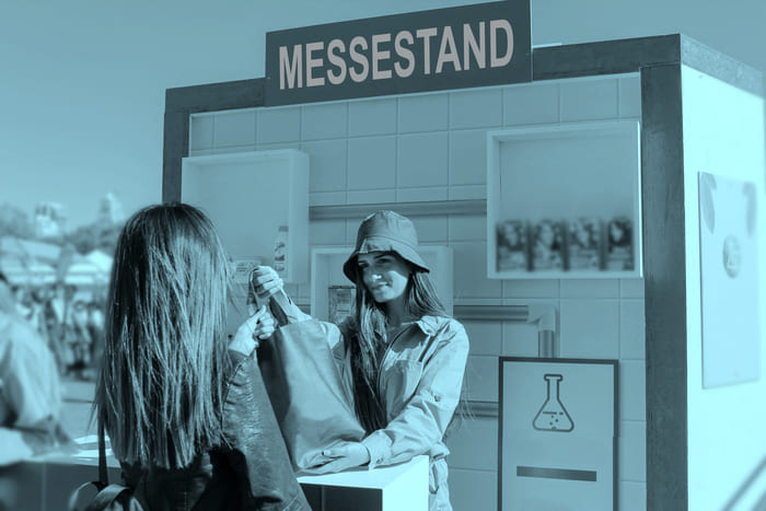 Messestand mit zwei Frauen, von denen eine der anderen ein Säckchen mit Werbematerial überreicht – steht für die Gewerbeversicherungen von RIANEVA, die Messestandversicherungen bieten.