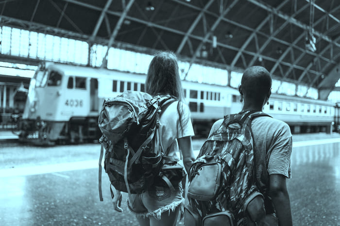 Zwei junge Frauen, eine davon schwarzer Herkunft, stehen mit Rucksäcken auf einem internationalen Bahnhof und gehen Richtung Zug – repräsentiert die RIANEVA Reiseschutzversicherung und die Werte der kulturellen Vielfalt.