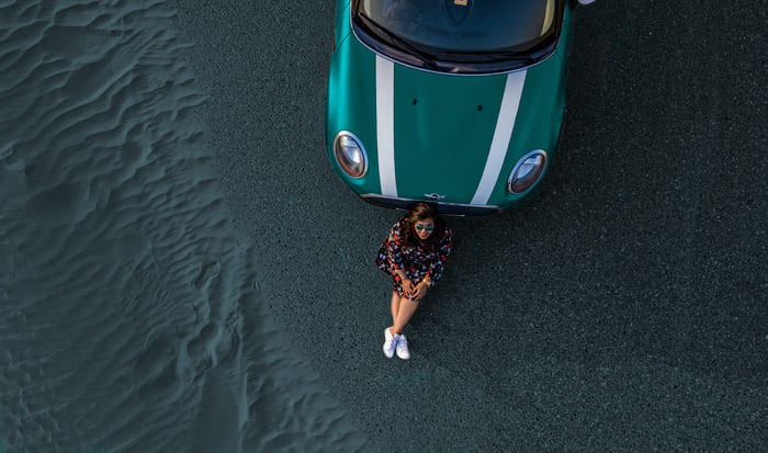 Eine Frau sitzt neben einem grünen Mini Cooper auf einer sandigen Fläche, trägt eine modische Kleidung und Sonnenbrille.